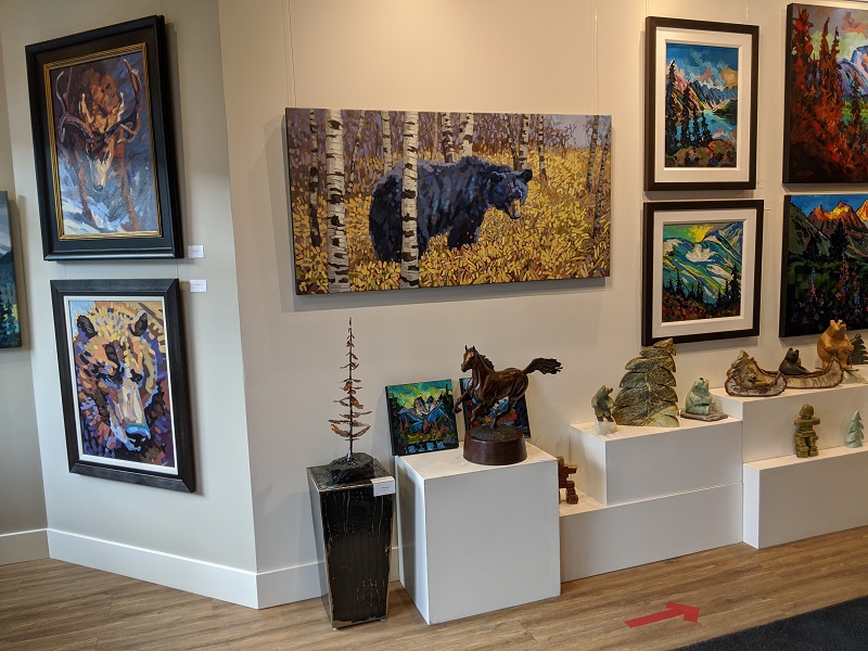 Interior of an art gallery with a painting of a black bear wandering up a forested hillside in the golden autumn. A painting of a stag with his head lowered and ready to battle, and a grizzly bear portrait painting are hung on the wall nearby.